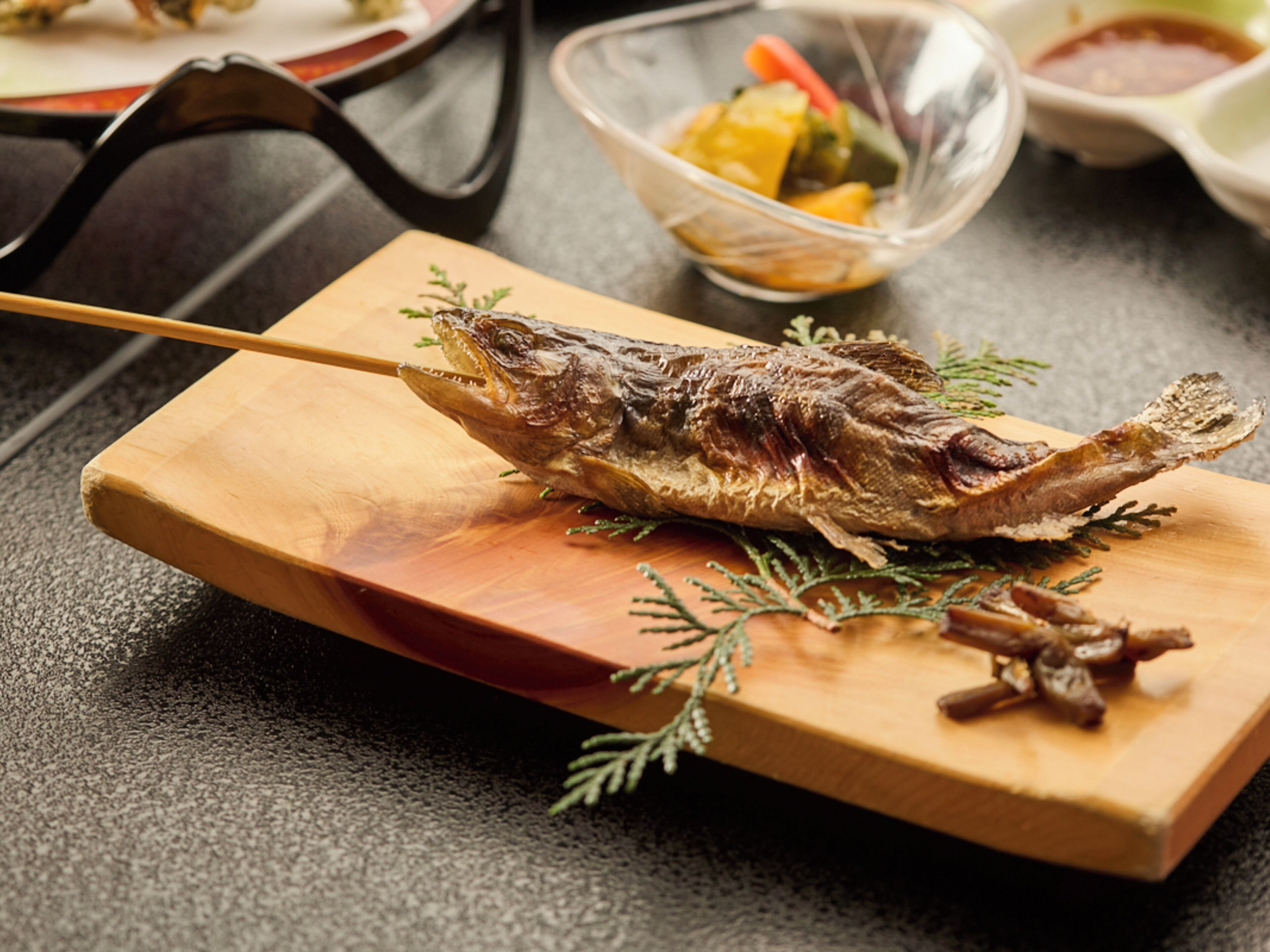 A grilled fish on a wooden board with a fork, surrounded by a bowl of vegetables and a small dish of sauce.