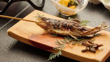 A grilled fish on a wooden board with a fork, surrounded by a bowl of vegetables and a small dish of sauce.