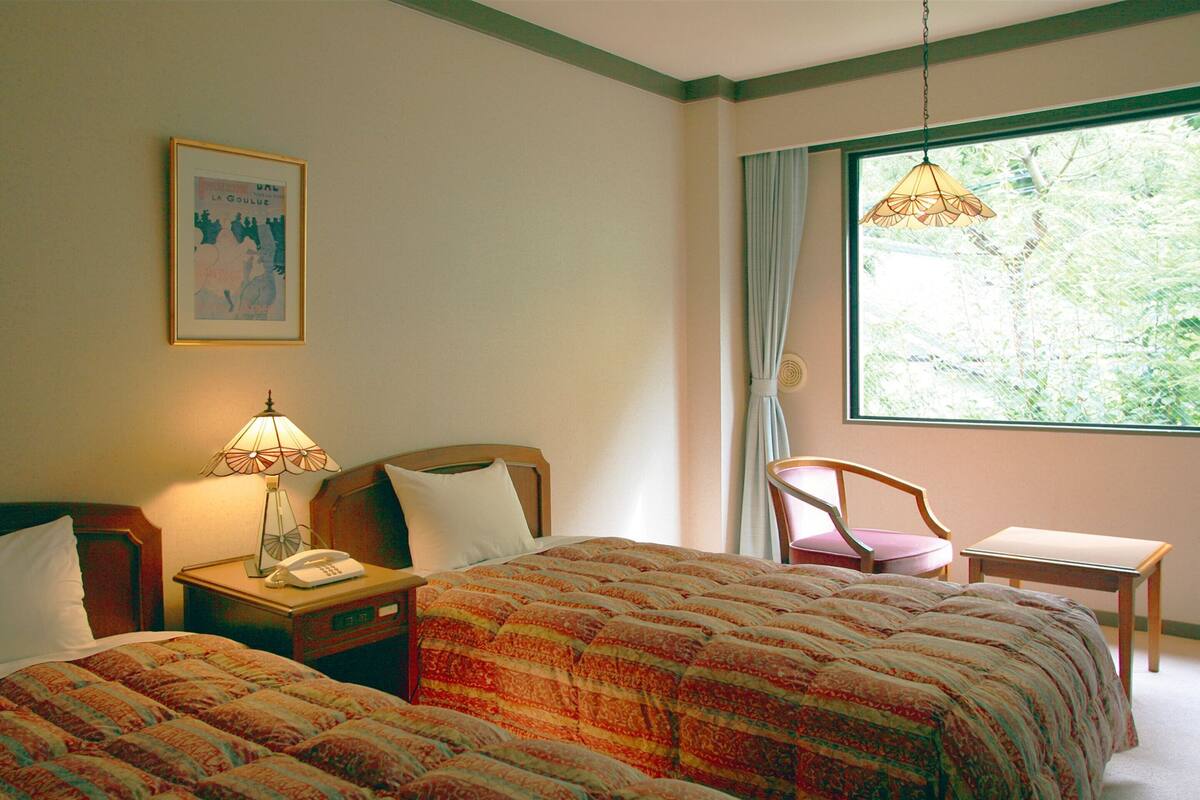 A hotel room with two beds, a desk, a chair, a lamp, a phone, and a window with a view of greenery.