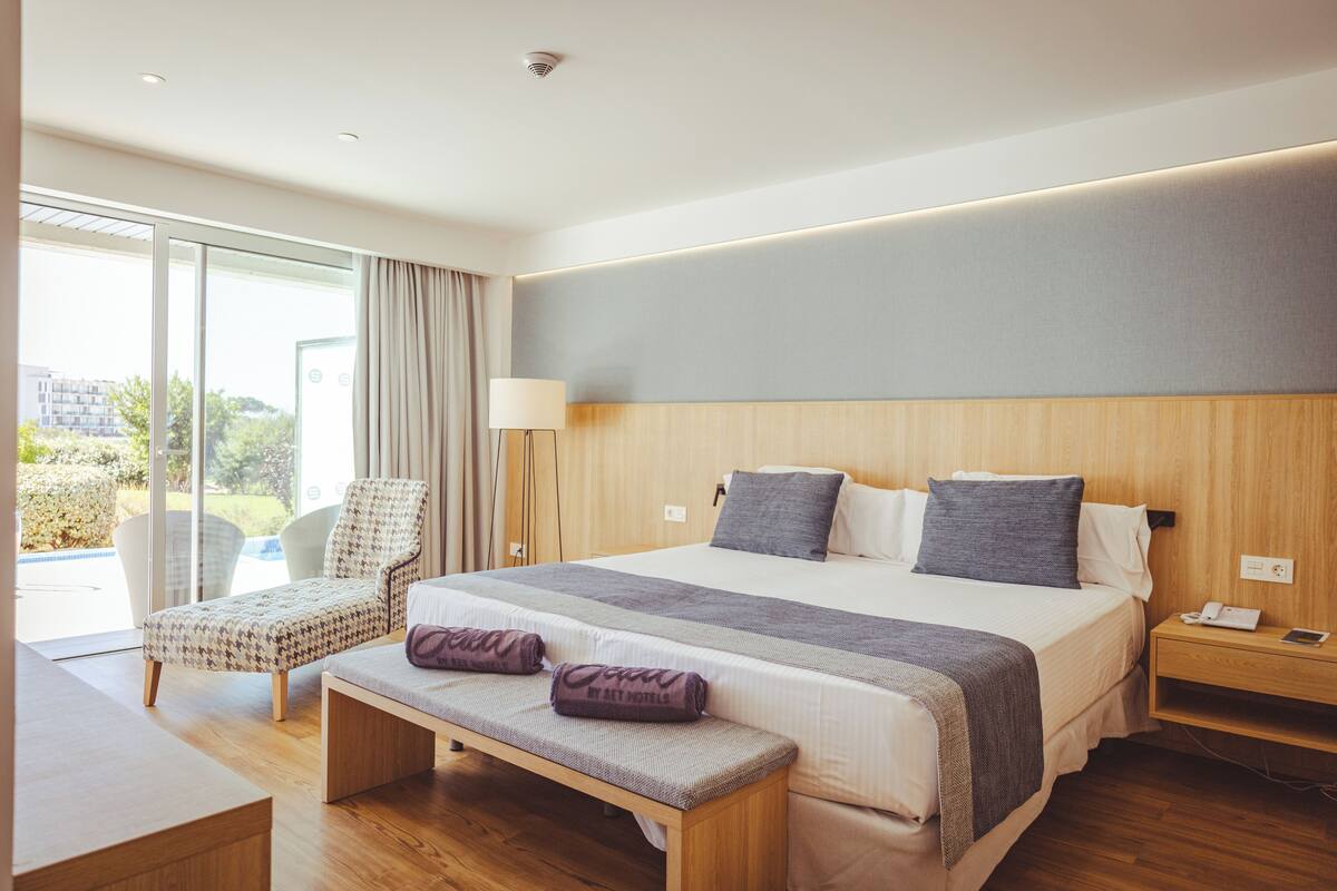 A modern hotel room with a large bed, a bench, a chair, a bedside table, and a view of the outdoors.