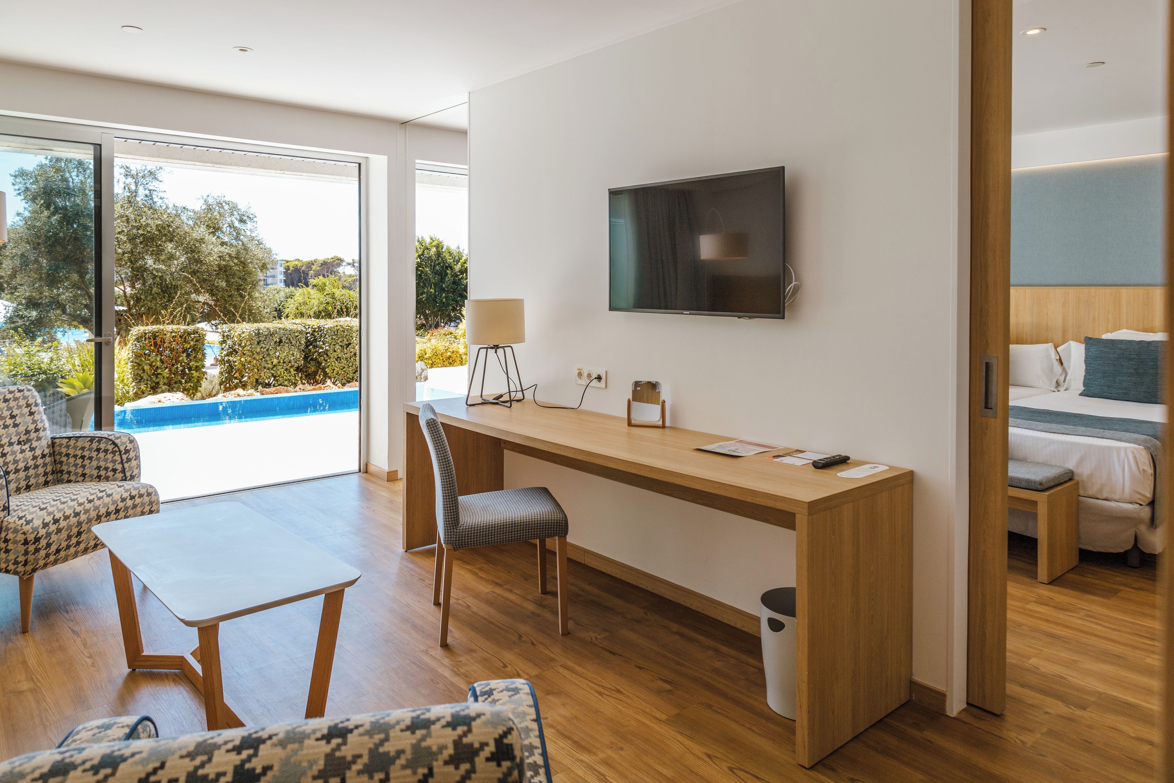 A modern hotel room with a wooden desk, a flat-screen TV, a sofa, and a view of a swimming pool.