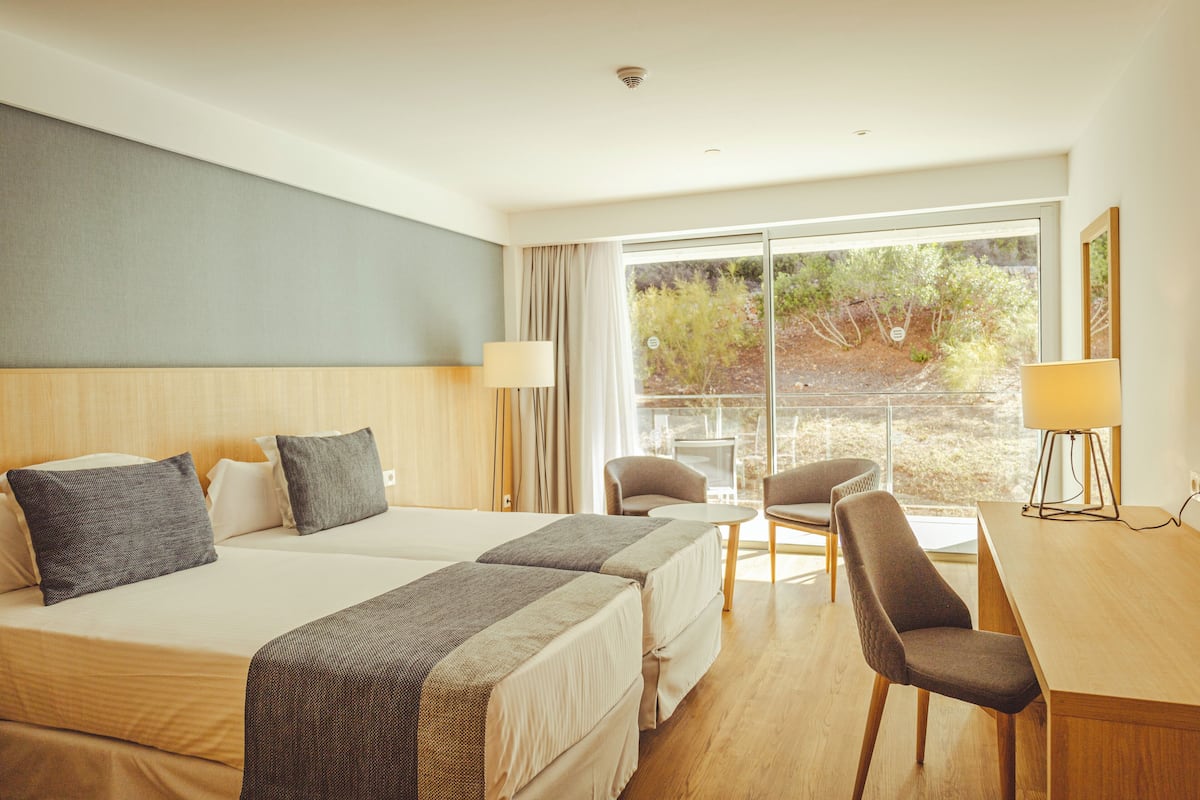 A hotel room with two beds, a desk, and a chair. There is a large window with a view of trees and a balcony.