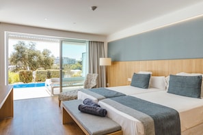 Quarto de hotel com cama, um banco, uma cadeira, um abajur e vista para uma piscina e vegetação lá fora.