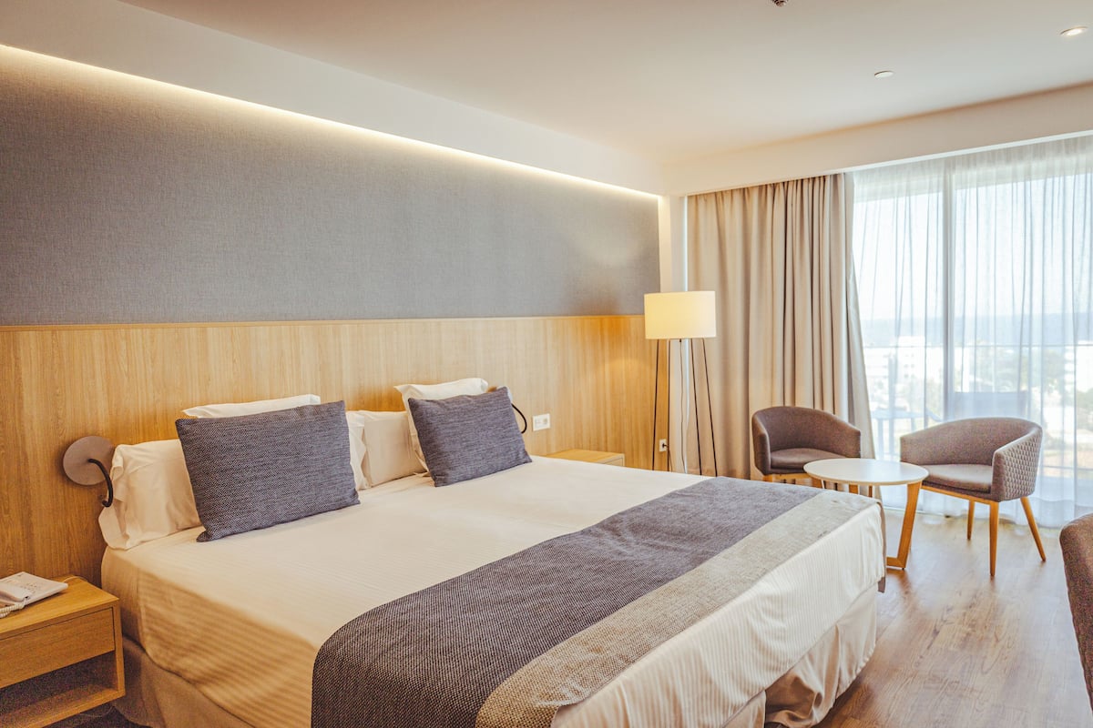 A hotel room with a large bed, two bedside tables, a desk, a chair, and a window with curtains.