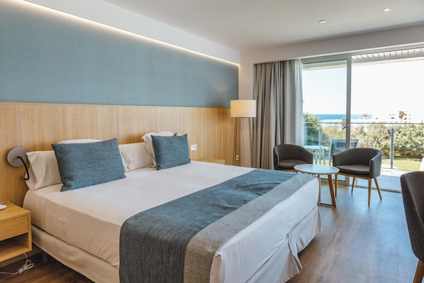 A hotel room with a large bed, a desk, a chair, a small table, and a view of the sea.