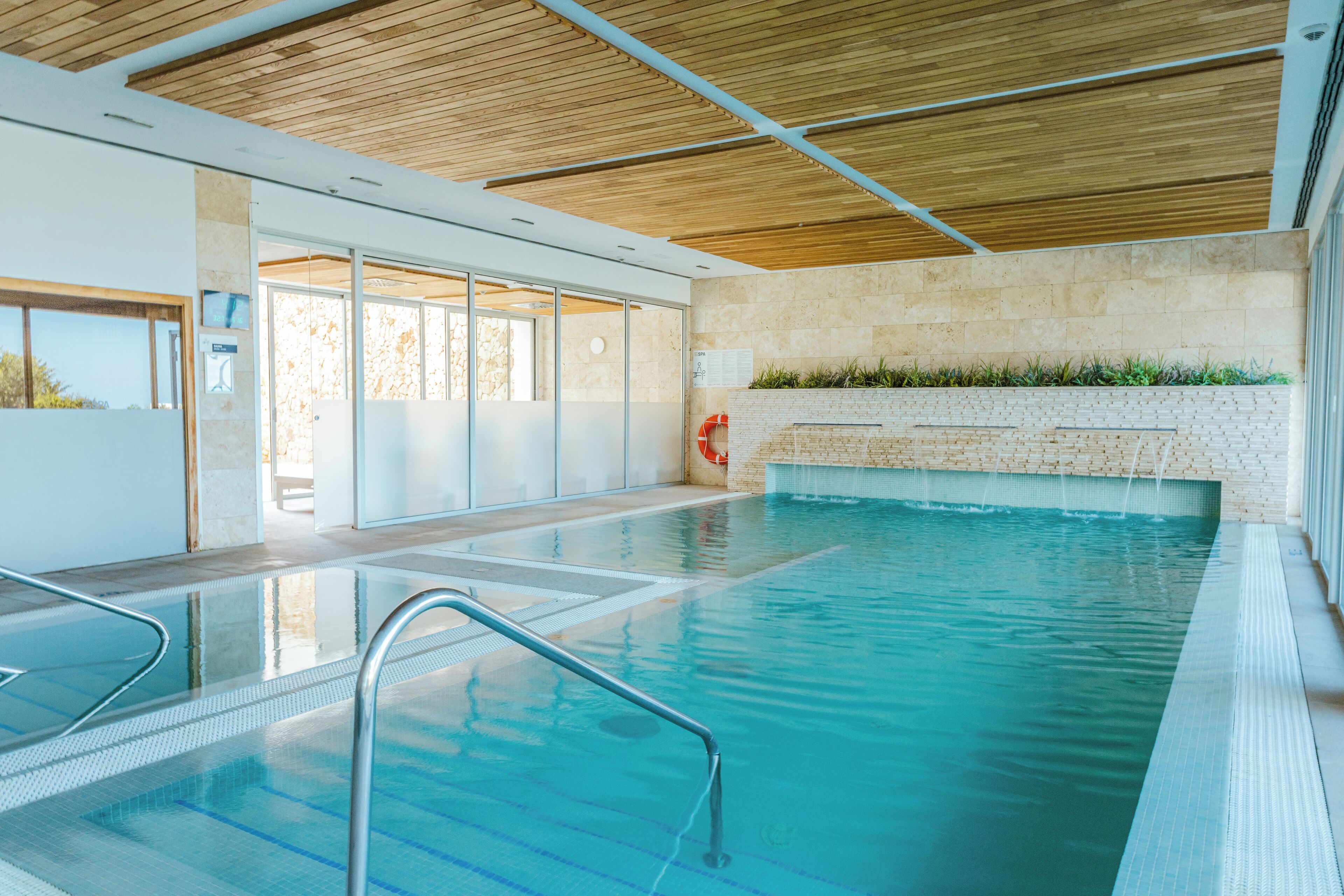 An indoor swimming pool with clear water, a wooden ceiling, and a brick wall with a plant border.