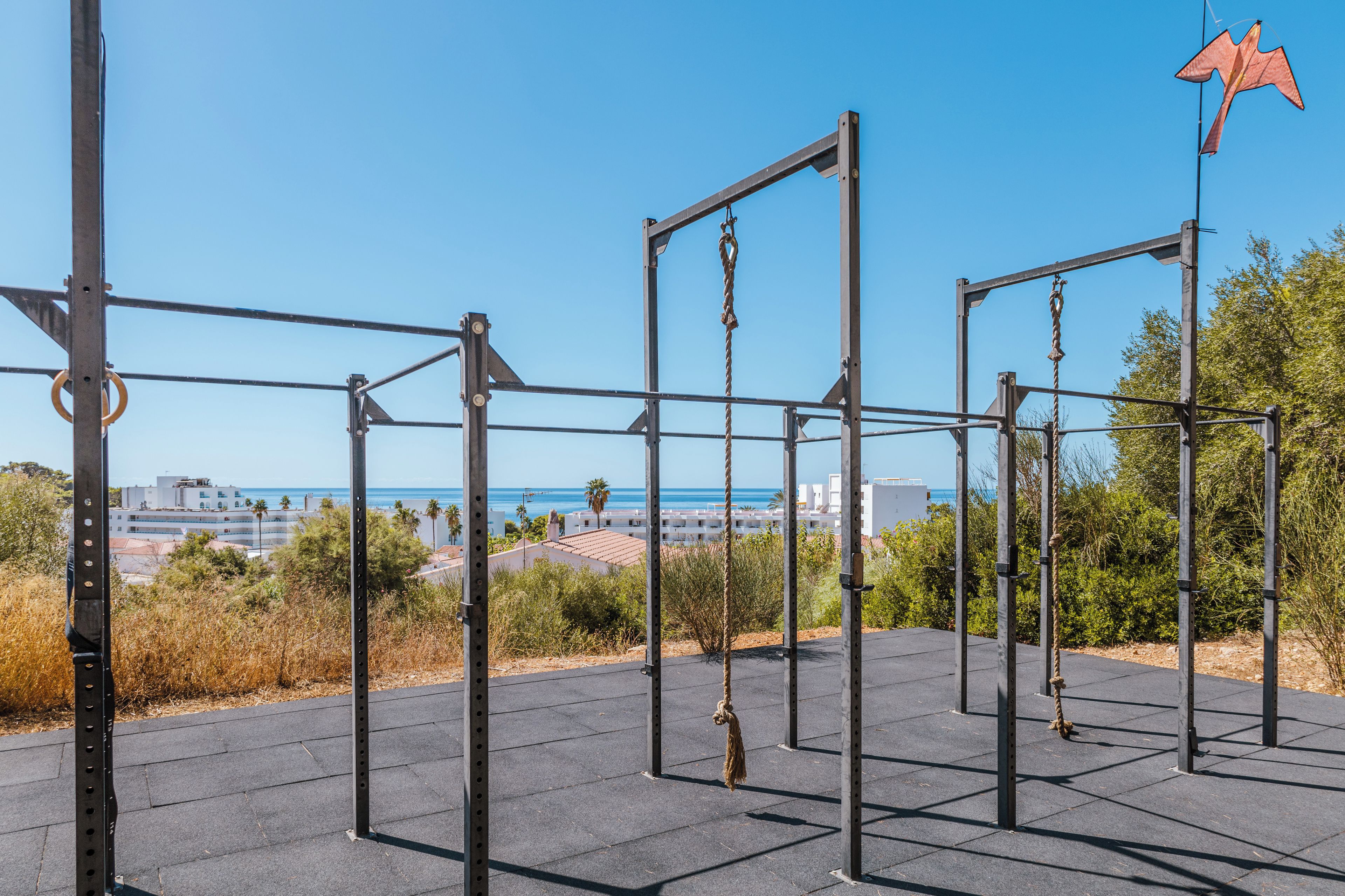 Un gimnasio al aire libre con estructuras metálicas y cuerdas, con vistas a una zona costera con edificios y el mar.