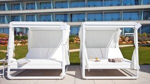 Two white outdoor lounge chairs with a table and a bottle of white wine between them, set against a modern building with multiple balconies.