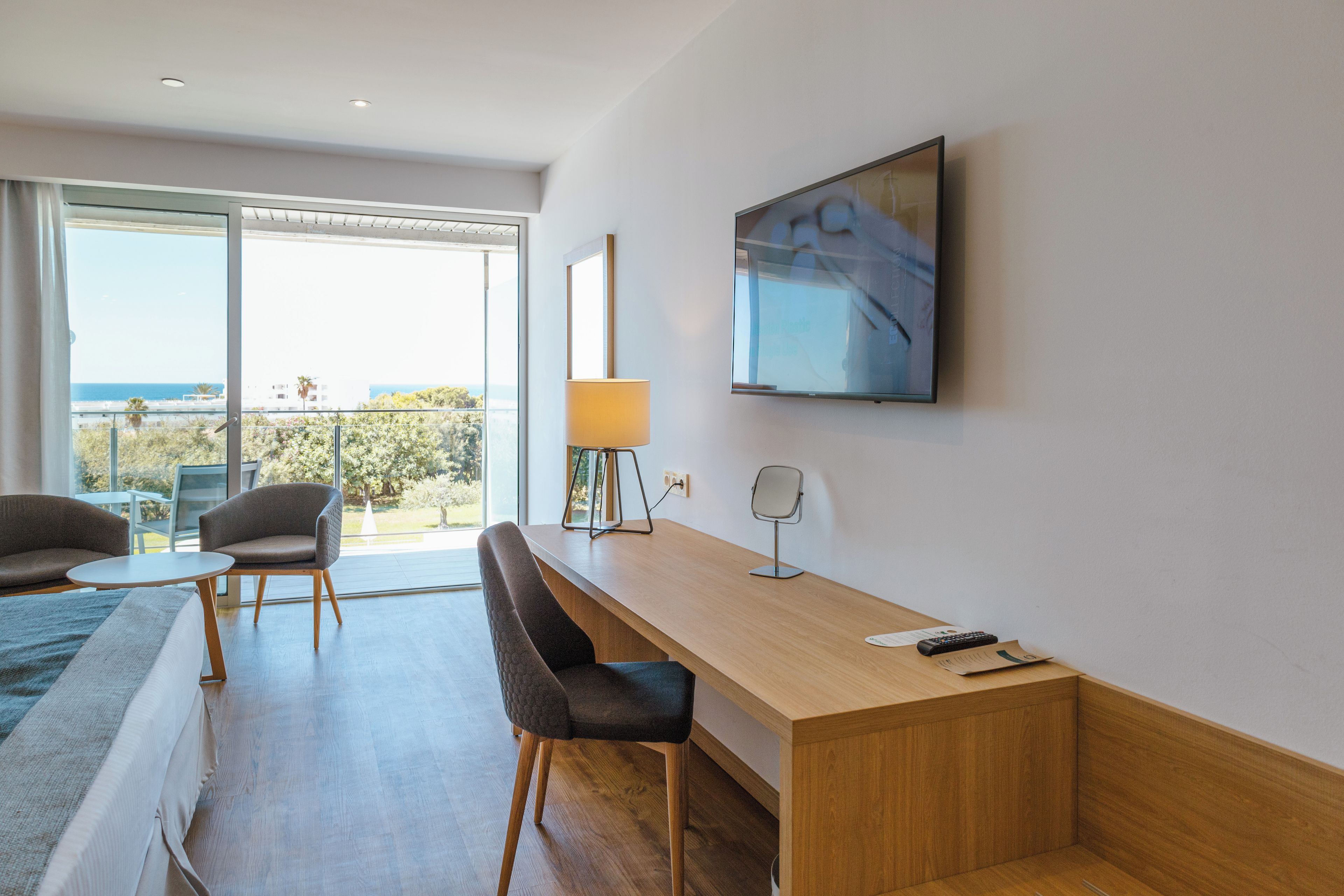 Habitación de hotel moderna con un ventanal grande, un escritorio, dos sillas, una lámpara y un televisor montado en la pared.
