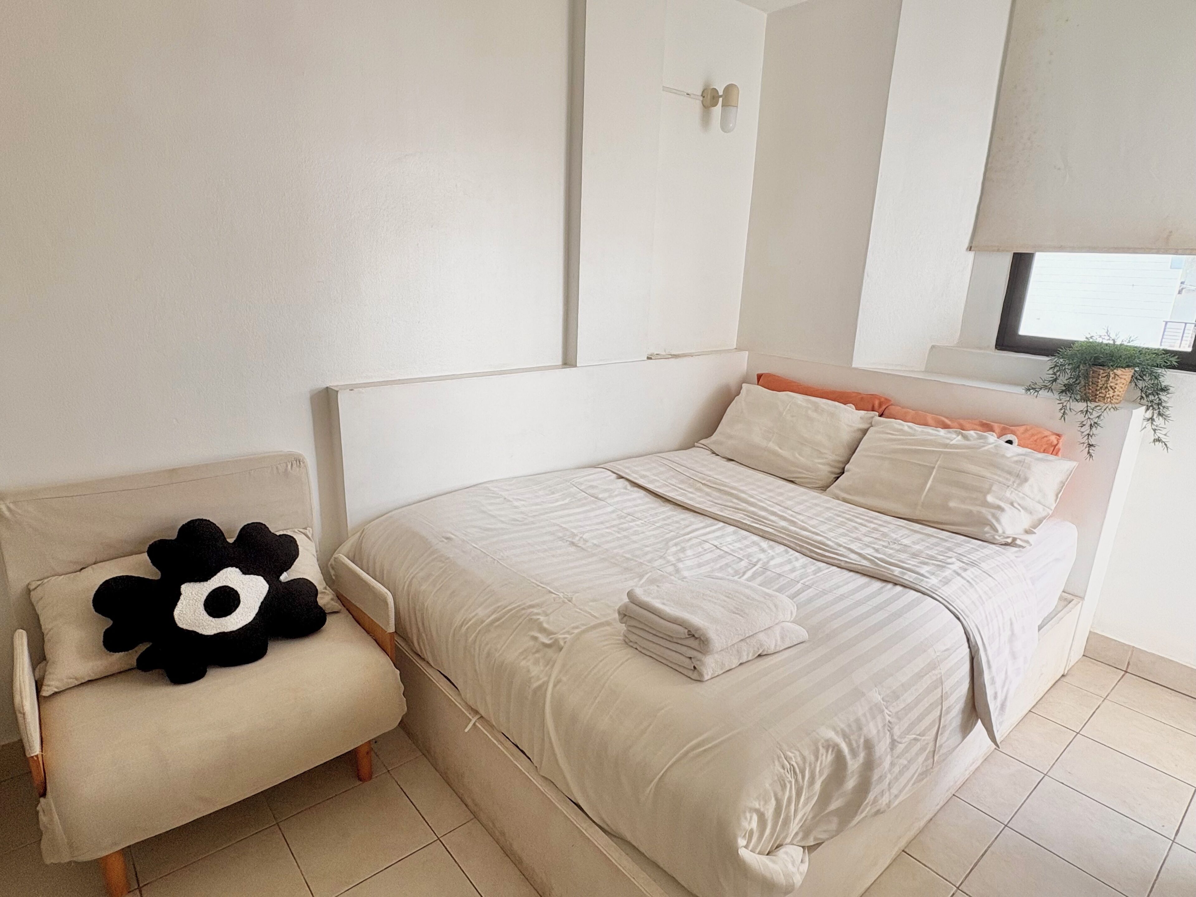 Un dormitorio ordenado con una cama, una silla y una ventana con una planta.