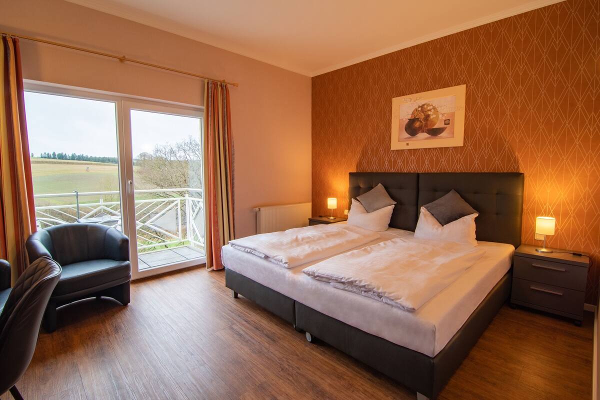 A hotel room with a large bed, a chair, a nightstand, and a view of a green landscape through the window.