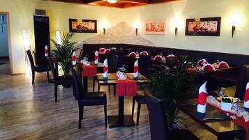 Un restaurante con suelo de madera, manteles rojos y sillas negras. Hay plantas en macetas y cuadros en las paredes.