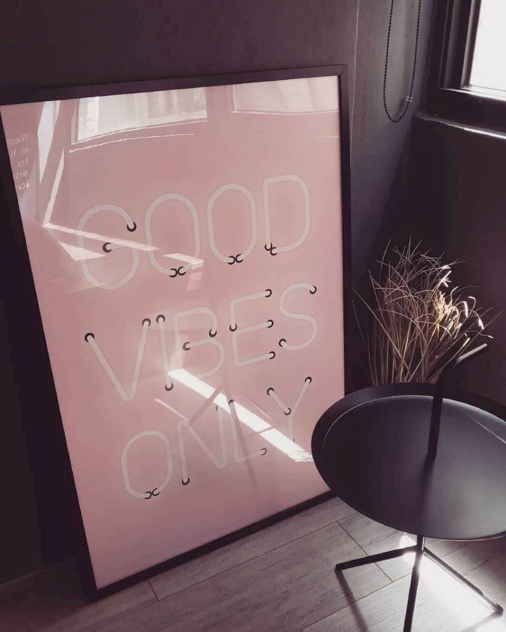 Una habitación con un letrero rosa grande, fijado a la pared, que dice 'GOOD VIBES ONLY' y una silla negra con un florero de plantas secas.