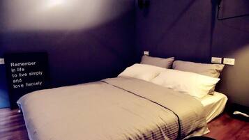 Una cama bien hecha con edredón y almohadas de color claro, una pared negra con un interruptor de luz, y una frase motivacional en la pared.
