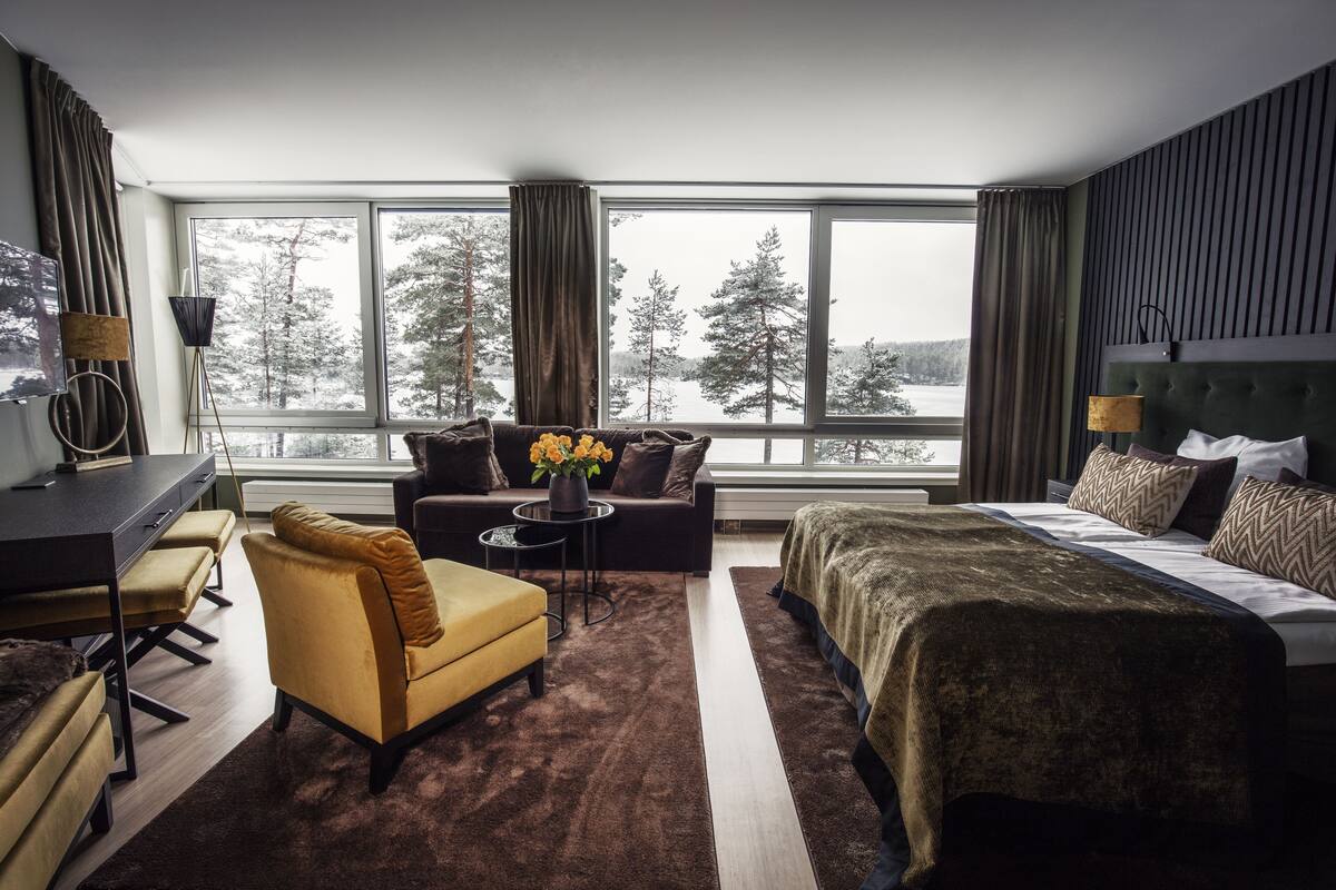 A modern hotel room with a large window, a sofa, a bed, a desk, and a chair.