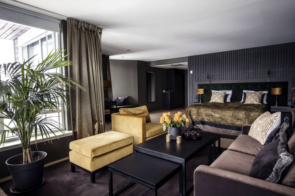 A modern hotel room with a large bed, a sofa, a coffee table, and a potted plant.