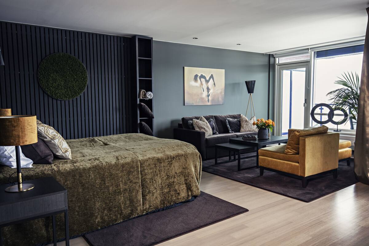 A modern bedroom with a bed, a sofa, a coffee table, and a chair. There is a window with a view of the outdoors.