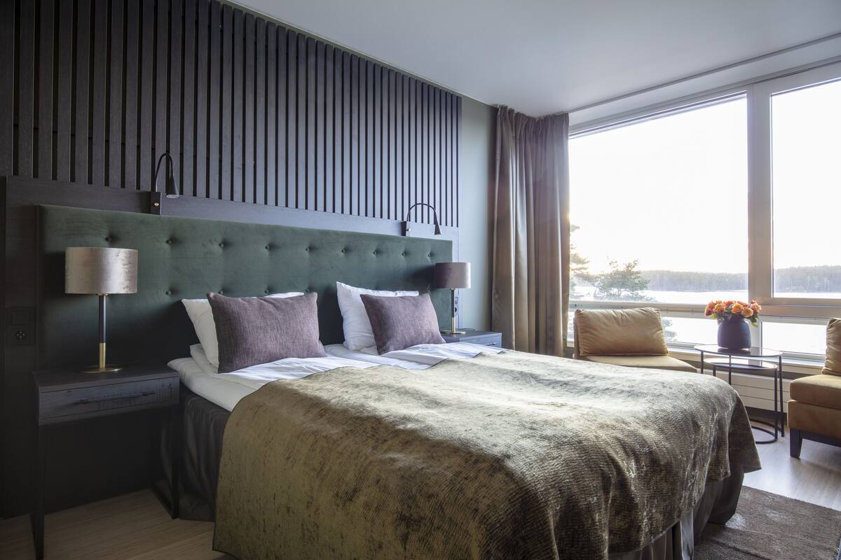 A modern hotel room with a large bed, a nightstand, a lamp, a chair, and a view of the outdoors through large windows.