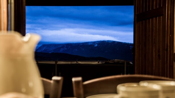 Blick auf ein Gebirge durch ein Fenster, im Vordergrund ein Tisch mit einer weißen Kanne und mehreren Tassen.