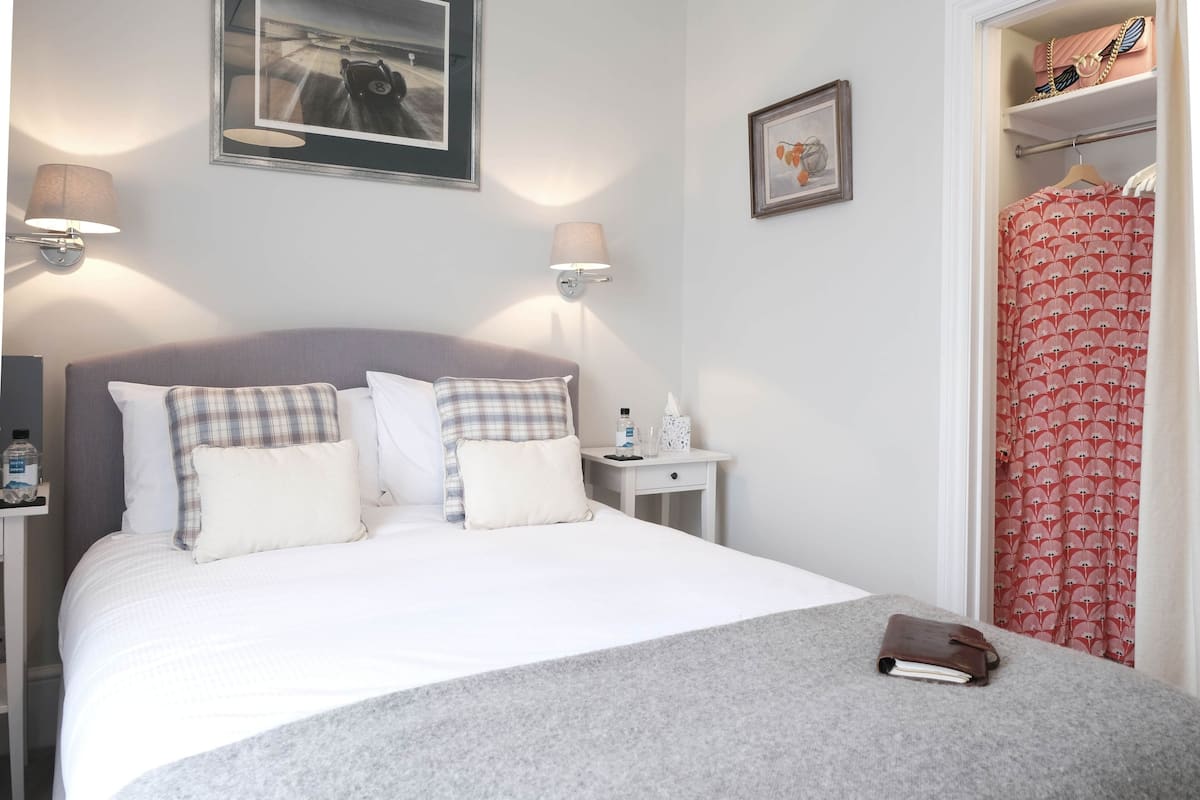 A neatly made bed with a grey blanket, a bedside table with a lamp, and a wardrobe with a red patterned curtain.