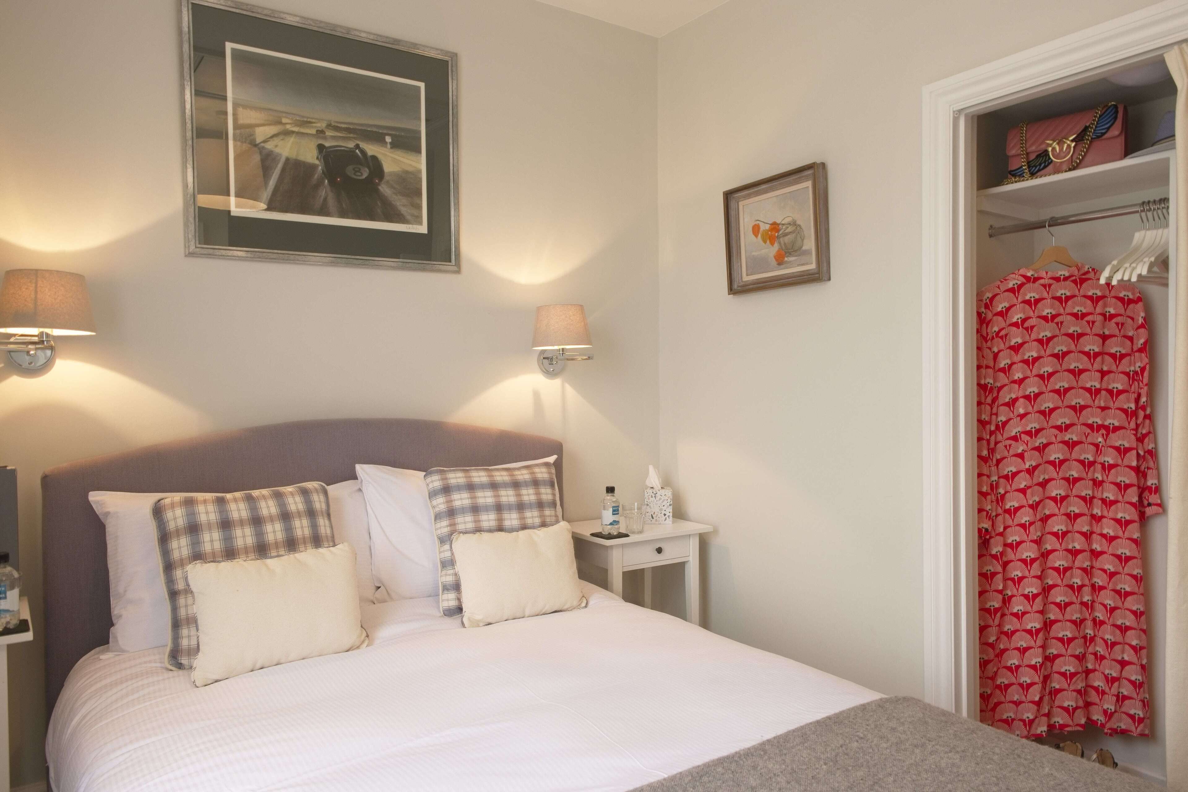 A neatly made bed with a headboard, two bedside tables with lamps, a framed picture on the wall, and a closet with a red patterned garment. - Saddlers (Chichester)