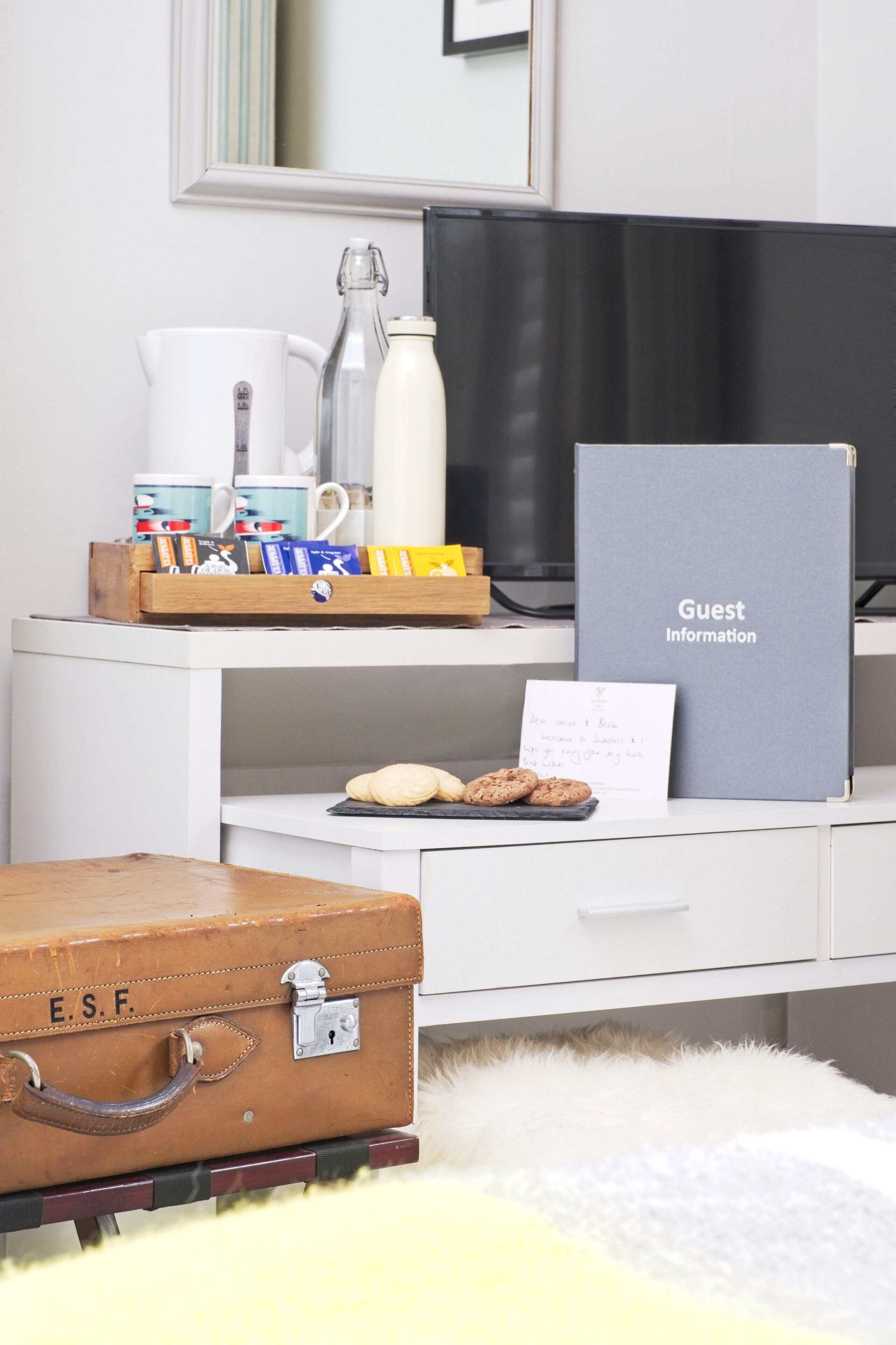 A neatly organized desk with a coffee maker, a bottle, a tray with books and snacks, a guest information folder, and a suitcase.