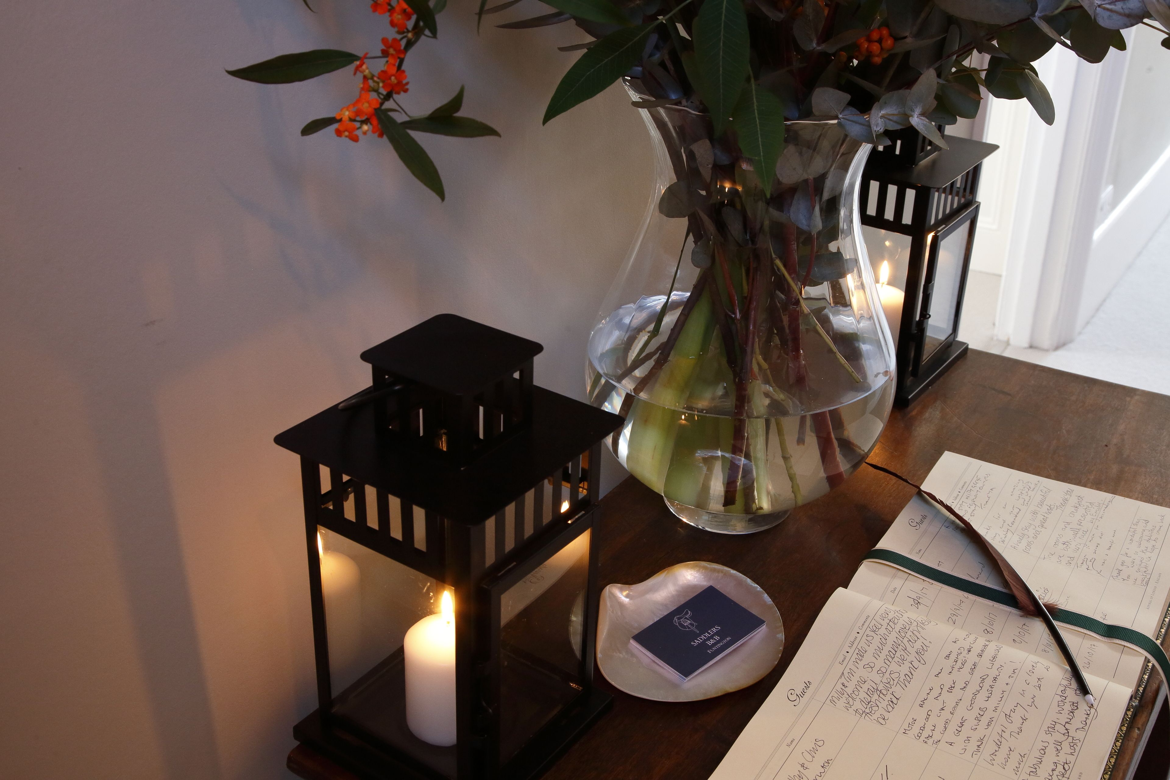 Una vela en un florero de vidrio, un farol negro y una libreta sobre una mesa de madera.