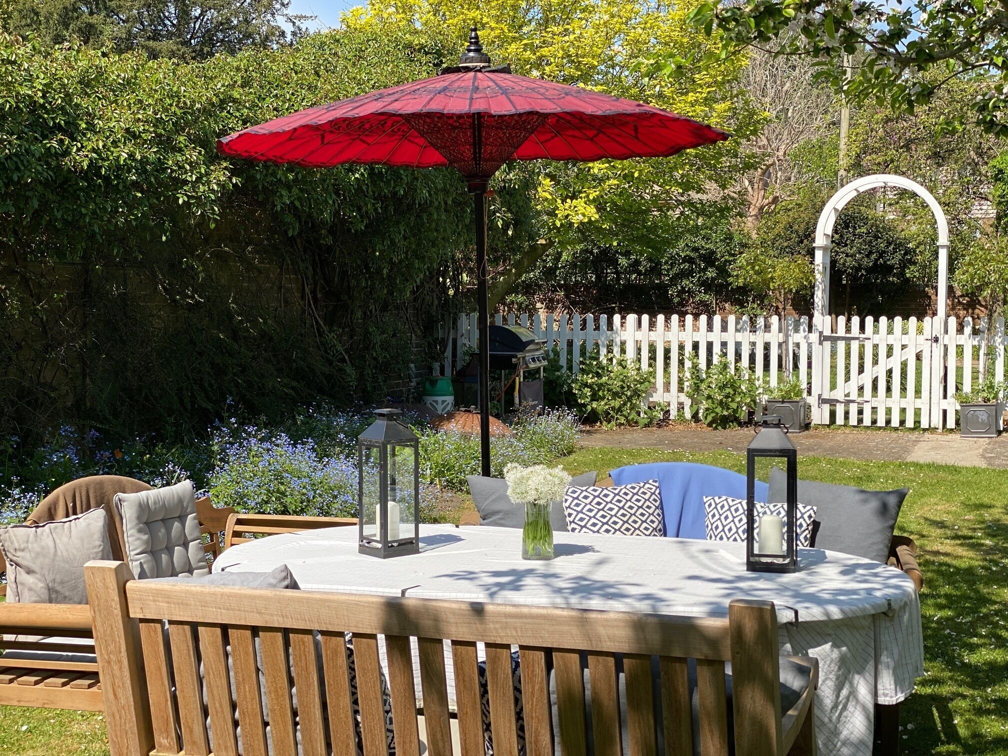 A round wooden table with a white tablecloth, surrounded by wooden chairs. There is a red umbrella providing shade, a lantern, and a vase with flowers on the table.