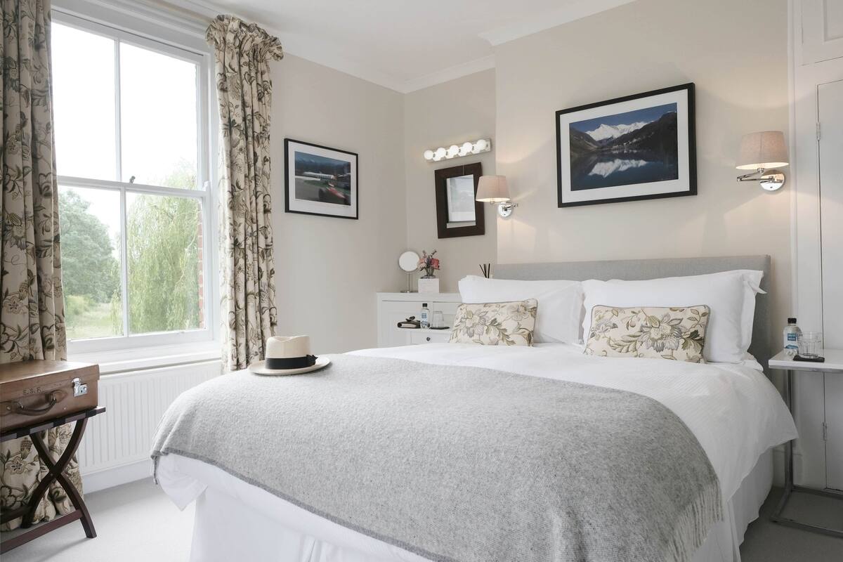 A neatly arranged bedroom with a large bed, a bedside table with a lamp, a suitcase, and a window with curtains.