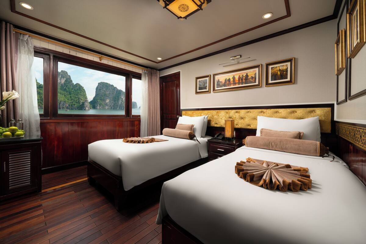 A hotel room with two beds, wooden flooring, a large window with a view of mountains, and framed pictures on the wall.