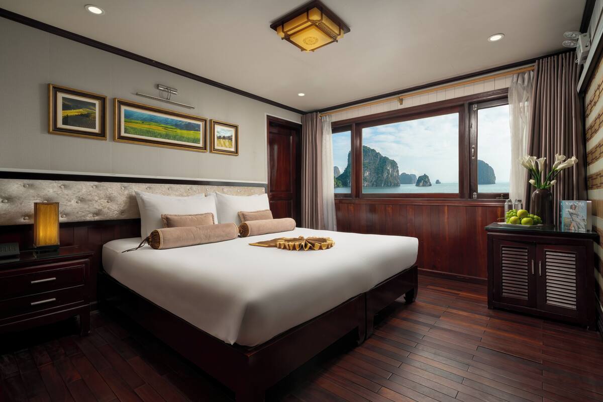 A spacious bedroom with a large bed, wooden flooring, a nightstand, a dresser, and a window offering a view of the sea and mountains.