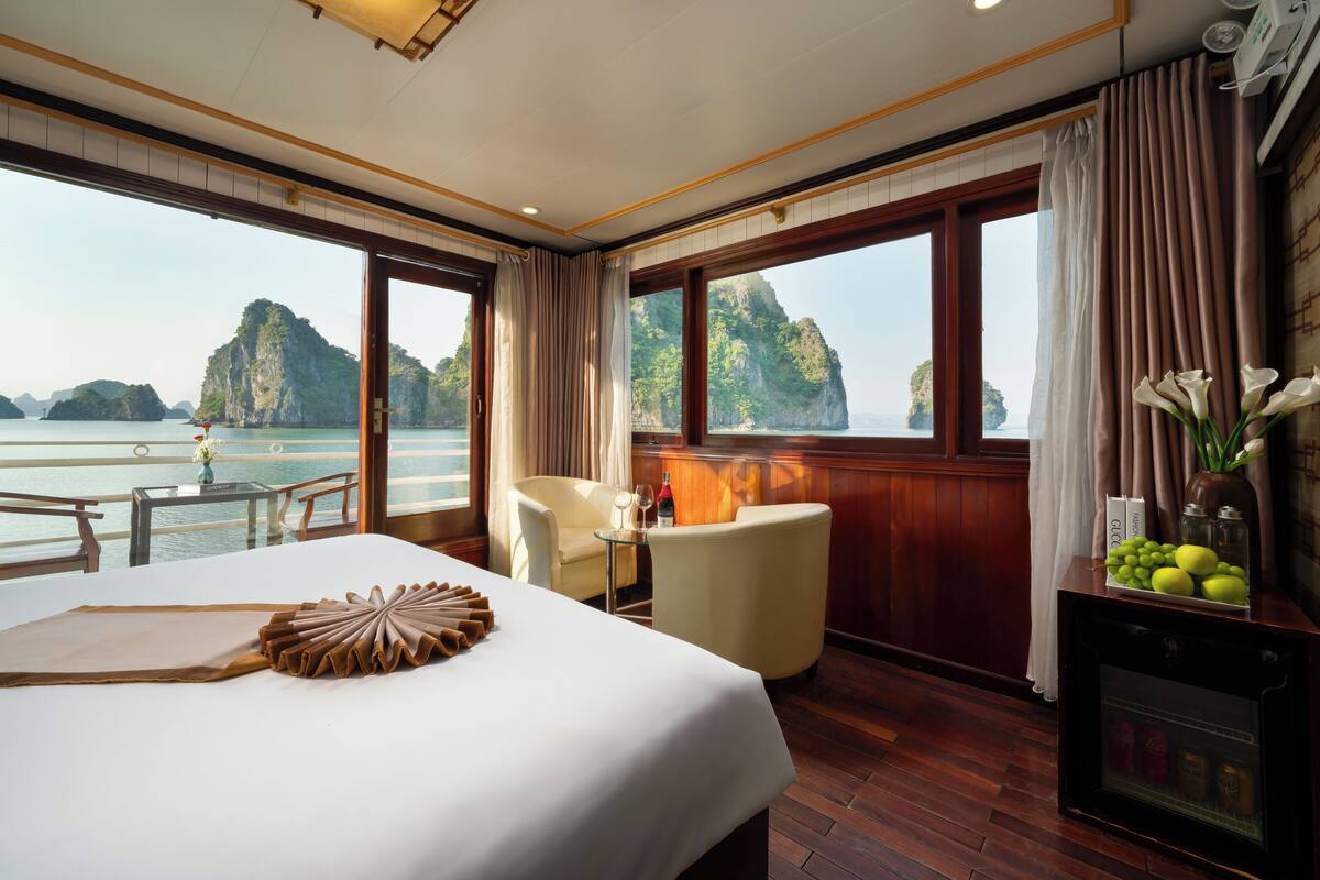 A hotel room with a large bed, a small table with a vase of flowers, a chair, and a view of the sea and mountains.