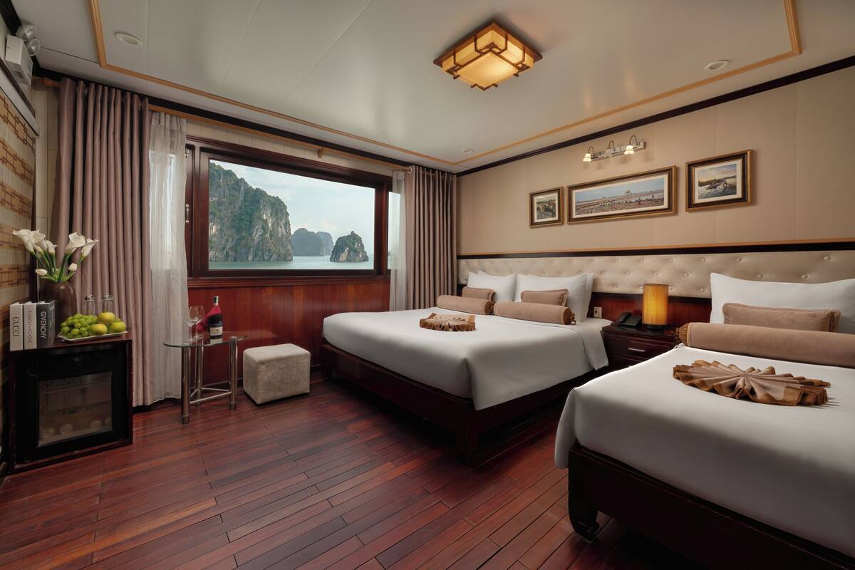 A hotel room with two beds, a wooden floor, a window with a view of cliffs, and a small table with a vase of flowers.