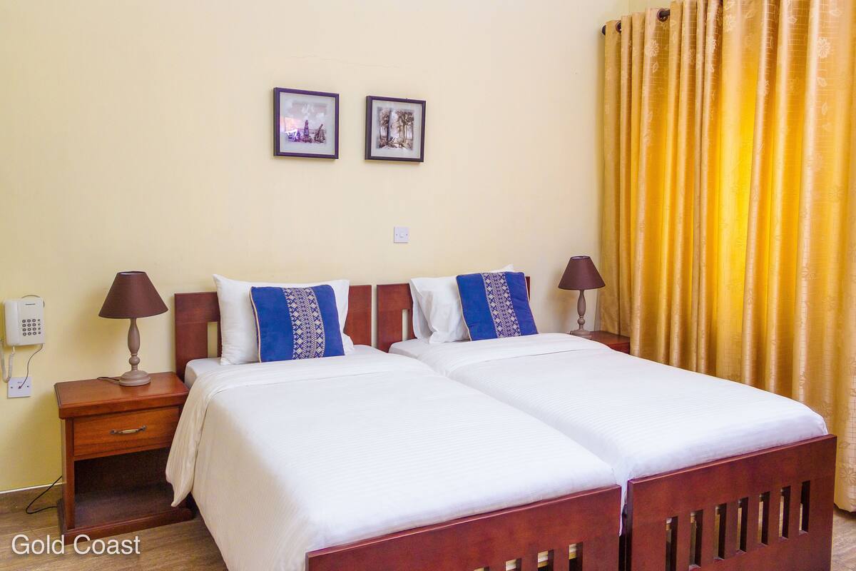 A double bed with white linens, two pillows, and a wooden headboard. There are two bedside tables with lamps, a wall-mounted telephone, and two framed pictures above the bed.