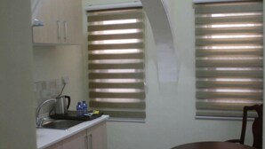 A compact kitchen with a sink, a kettle, and a small table with chairs. There are two windows with blinds, and the room has a tiled floor. - Lake Bosumtwi Hotel & Apartments (Accra)