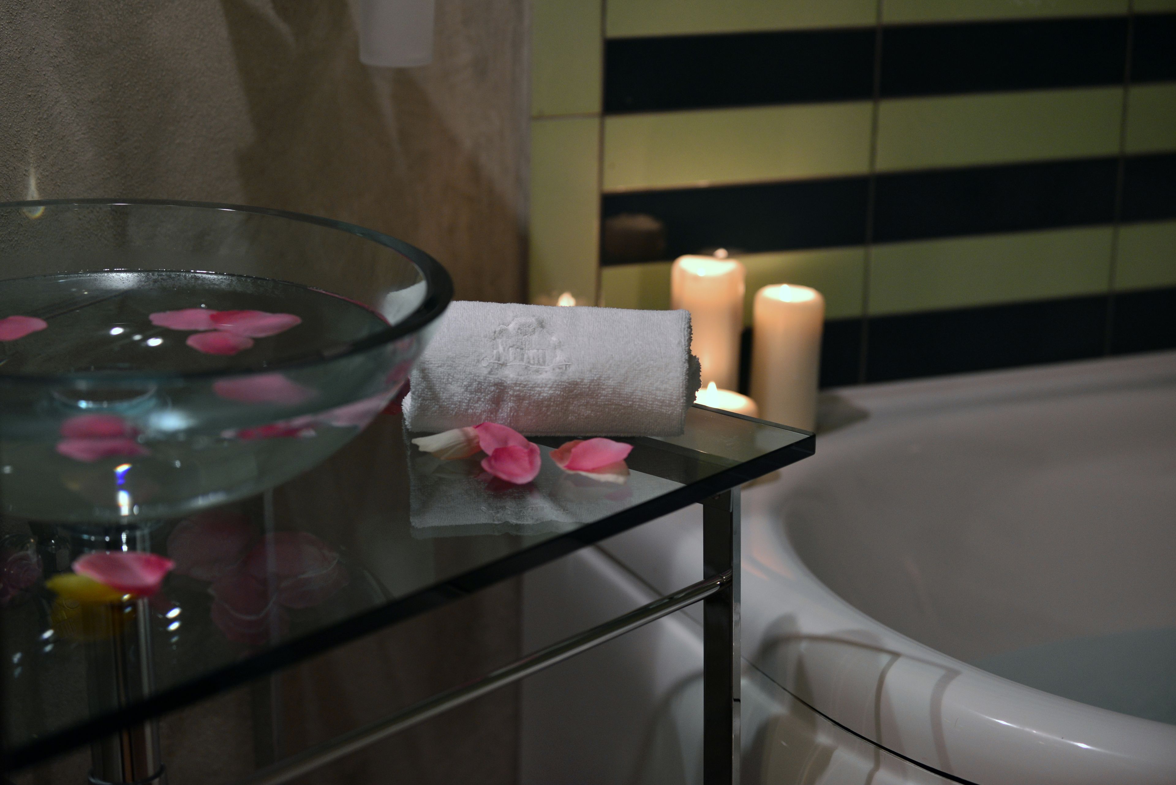 A glass bowl with rose petals and a towel on a glass table in a bathroom with a bathtub and candles.