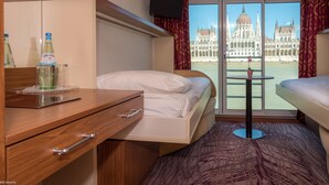 A hotel room with a bed, a desk with a bottle and glasses, a TV, and a view of a large building through the window. - Select MS Serenity - Neuss (Neuss)