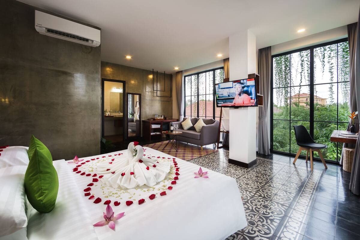 A hotel room with a neatly made bed, a TV, a sofa, and a dining area with a view of greenery.