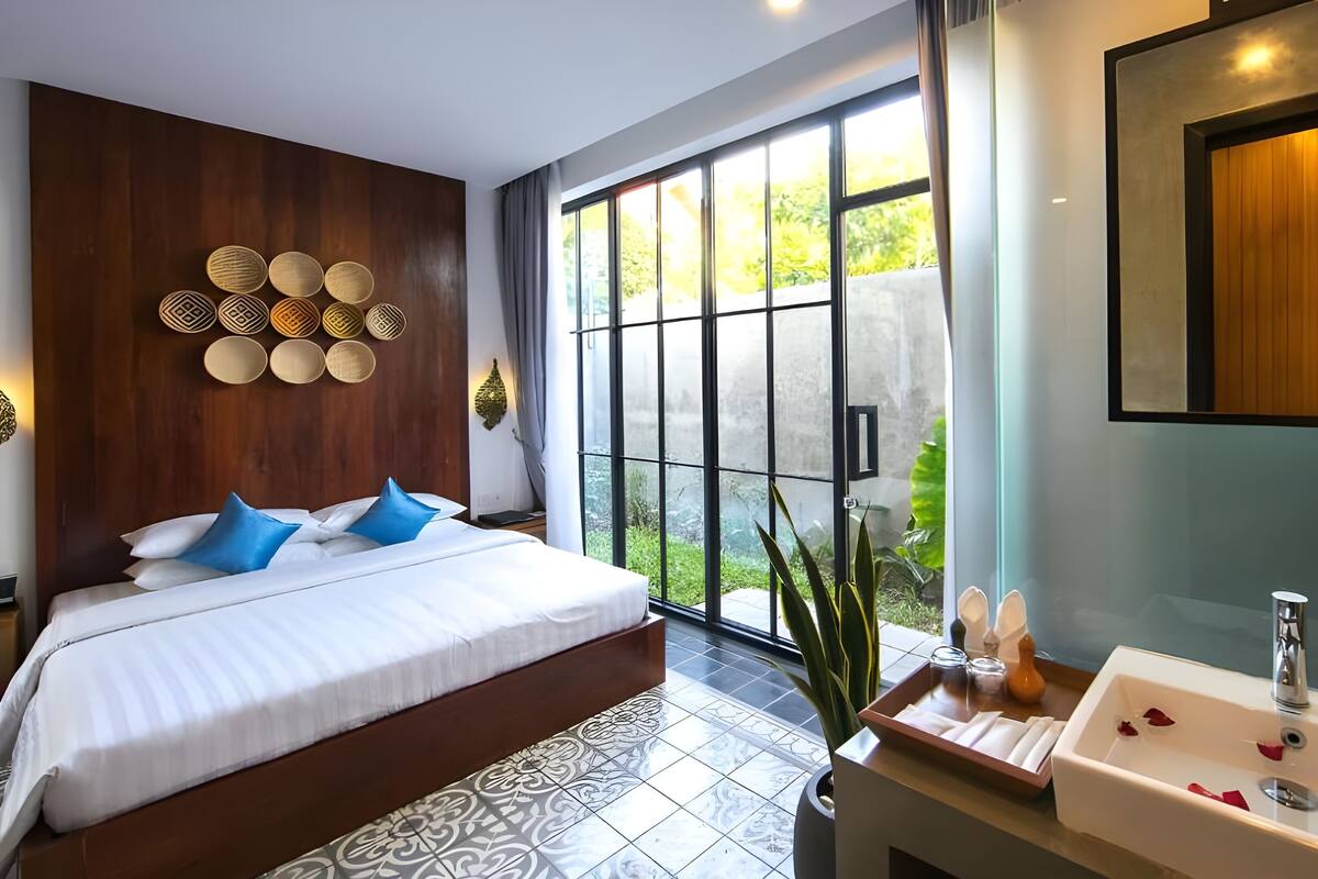 A modern bedroom with a large bed, a bathroom with a sink and shower, and a view of greenery through large windows.