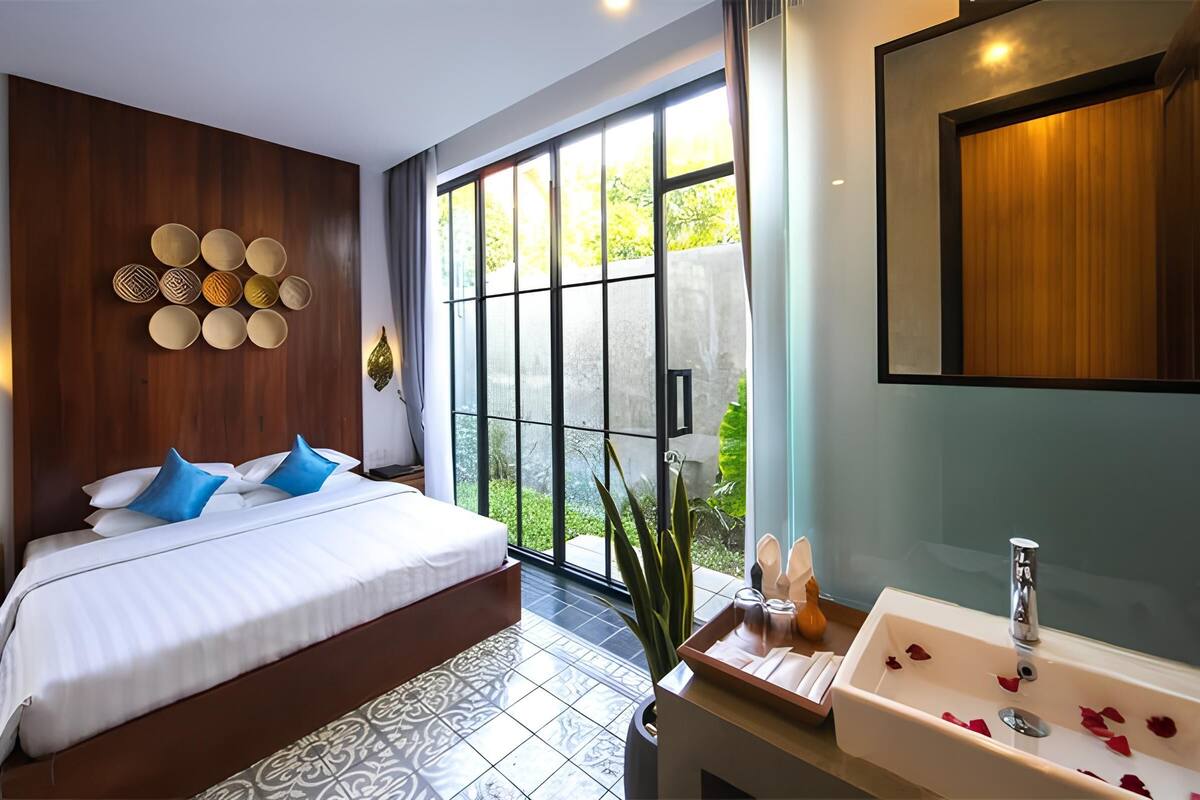 A modern hotel room with a large bed, a bathroom with a sink and mirror, and a view of greenery through large windows.