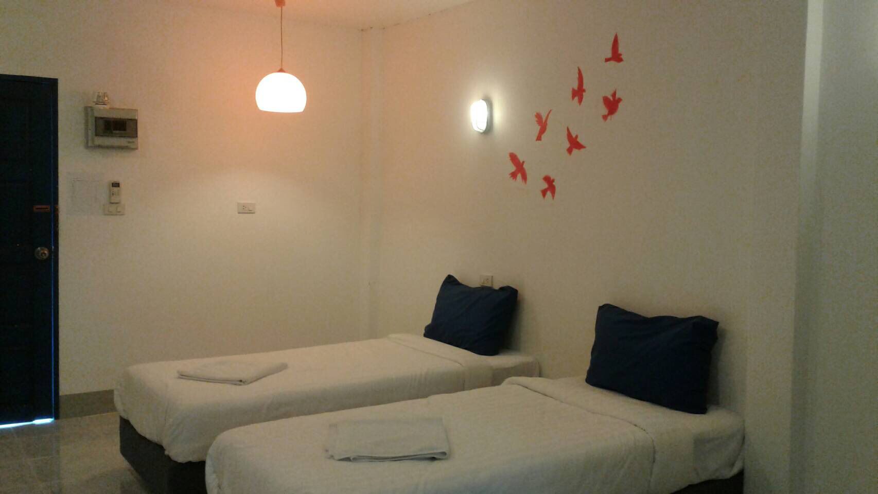 A room with two single beds, each with a pillow and a folded towel. There is a wall-mounted light fixture above the beds and a wall-mounted air conditioning unit.