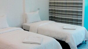 A hotel room with two single beds, each with white linens and pillows, a striped curtain, and a small bedside table with a book. - Blue Hip Apartment Nakhon Sri (Nakhon Si Thammarat)