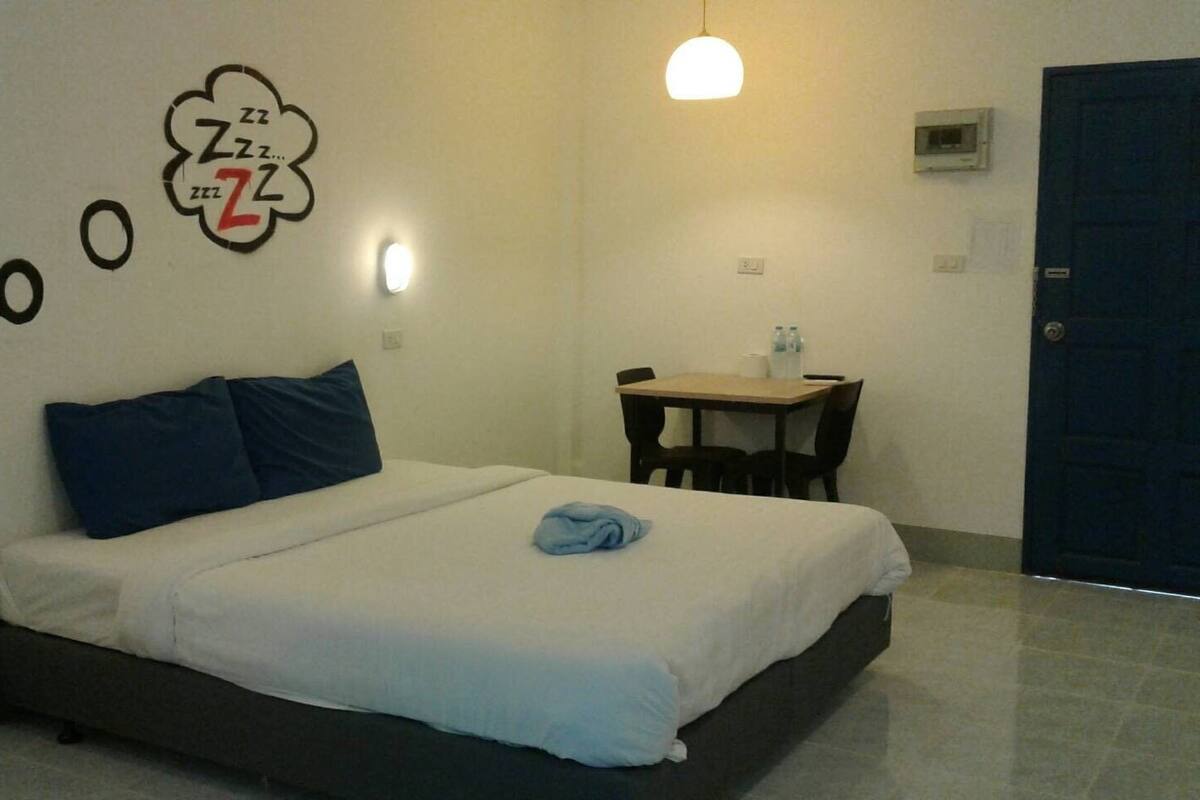 A single bed with a white mattress and dark headboard, a small table with chairs, a wall clock, and a door.