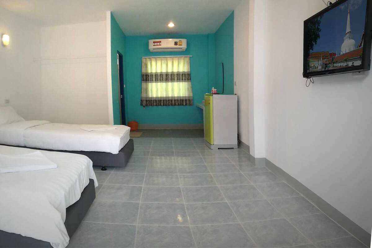 A hotel room with two beds, a small kitchenette, a window with curtains, and a wall-mounted TV.