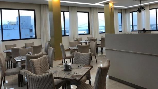 A modern hotel dining area with multiple tables and chairs, large windows with curtains, and a view of the cityscape.