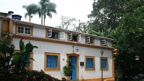 A two-story building with a white facade, blue door, and yellow trim. There are multiple windows with blue shutters, a small garden area with flowers, and a street lamp. - Pousada Richard Rothe (Tiradentes)