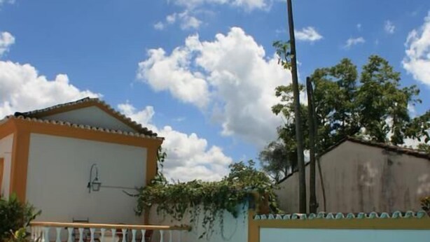 A swimming pool with a tiled deck, a building with a balcony, and a clear blue sky with clouds. - Pousada Richard Rothe (Tiradentes)