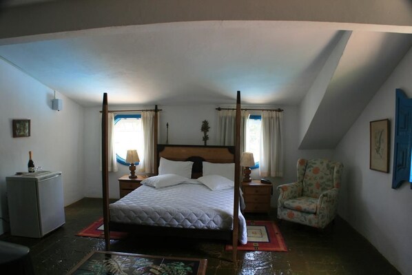 Un lit à baldaquin dans une pièce dont le plafond est en pente, avec un petit bureau, une chaise et un tableau encadré au mur.