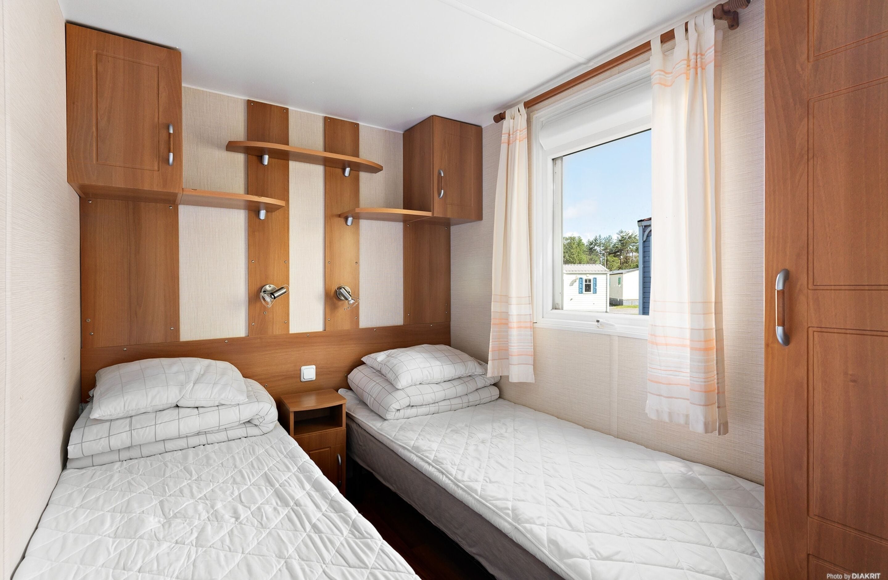 A compact bedroom with two beds, wooden cabinets, a small bedside table, and a window with curtains.