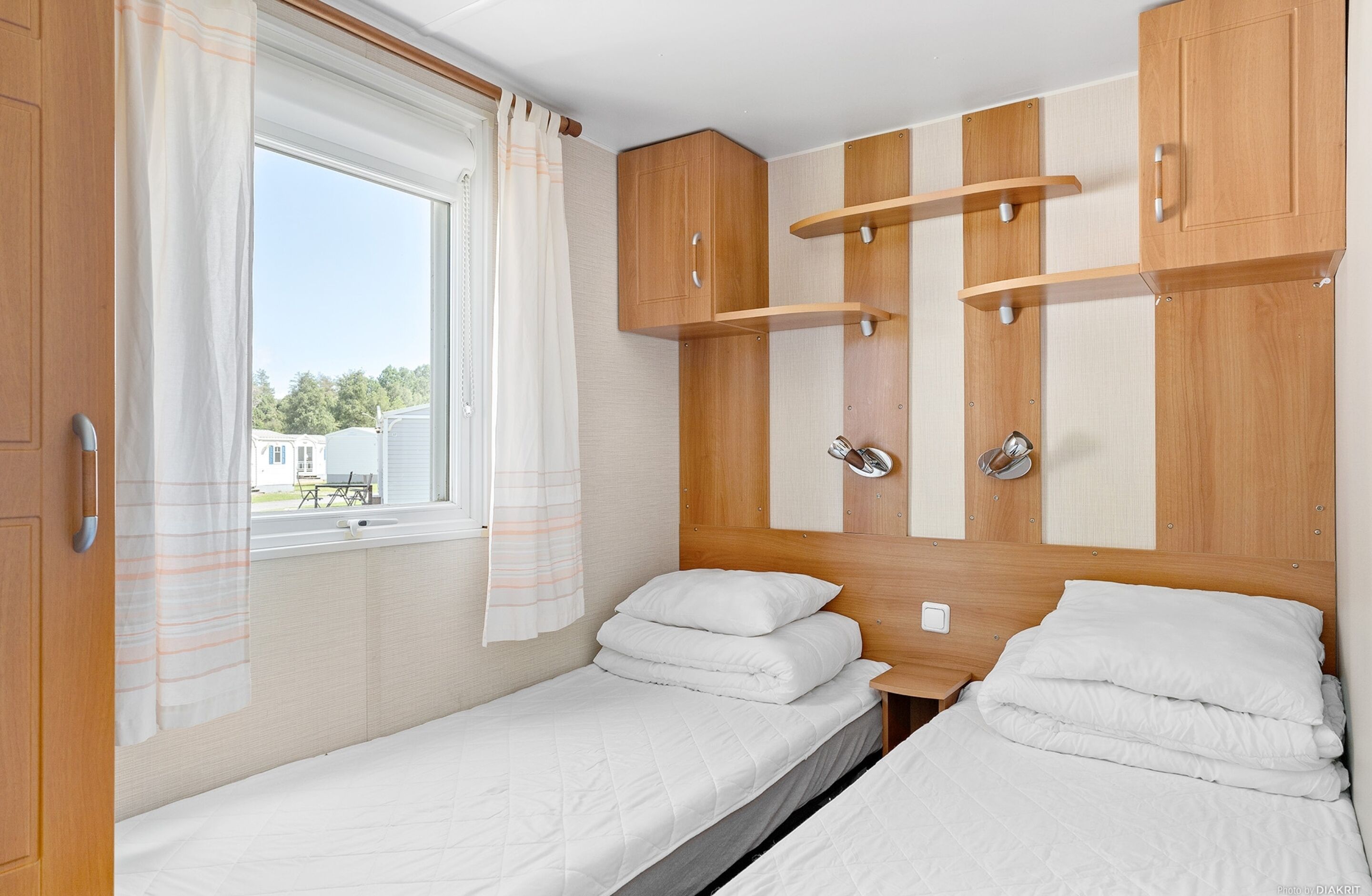 Un dormitorio pequeño con dos camas individuales, armarios de madera, una ventana con cortinas y vista a una casa afuera.